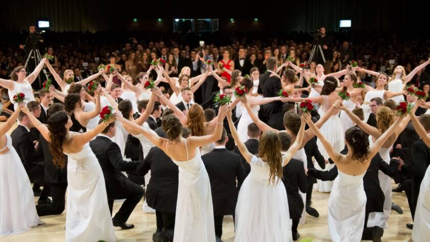 Eine große Gruppe formell gekleideter Frauen in weißen Kleidern und Männer in schwarzen Anzügen tanzen auf einem Maturaball einen koordinierten Tanz, bei dem sie sich an den Händen halten und das Publikum bei dieser großen Indoor-Veranstaltung in ihren Bann ziehen.