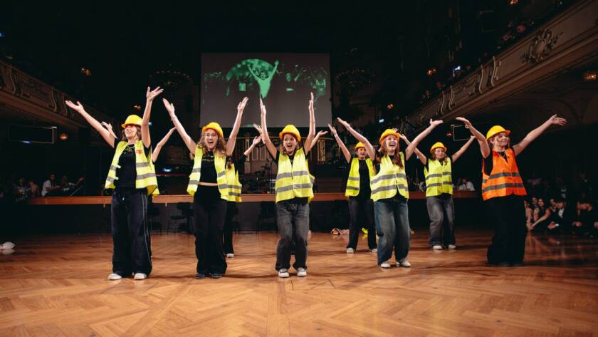 Eine Gruppe von Personen mit gelben Sicherheitswesten und Schutzhelmen führt einen choreografierten Tanz auf einer Holzbühne auf und hebt dabei die Arme. Im Hintergrund sind eine große Leinwand und das Publikum zu sehen.