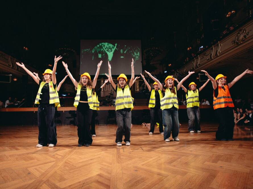 Eine Gruppe von Personen mit gelben Sicherheitswesten und Schutzhelmen führt einen choreografierten Tanz auf einer Holzbühne auf und hebt dabei die Arme. Im Hintergrund sind eine große Leinwand und das Publikum zu sehen.