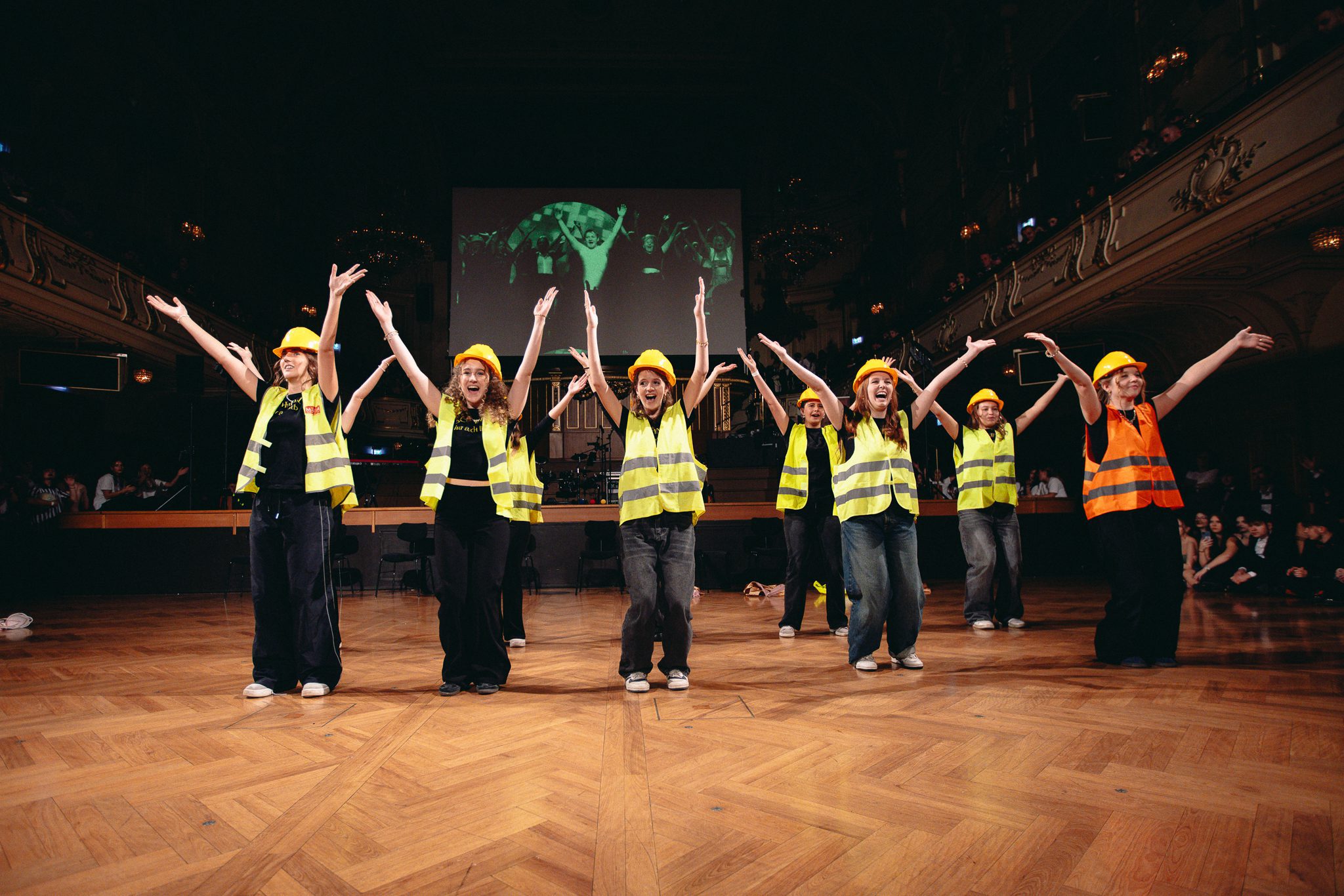 Eine Gruppe von Personen mit gelben Sicherheitswesten und Schutzhelmen führt einen choreografierten Tanz auf einer Holzbühne auf und hebt dabei die Arme. Im Hintergrund sind eine große Leinwand und das Publikum zu sehen.
