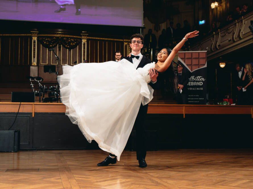 Ein Mann im Smoking hebt eine Frau in einem weißen Ballkleid hoch, während er in einem großen Ballsaal tanzt, während das Publikum von Balkonen und von der Seite zusieht.