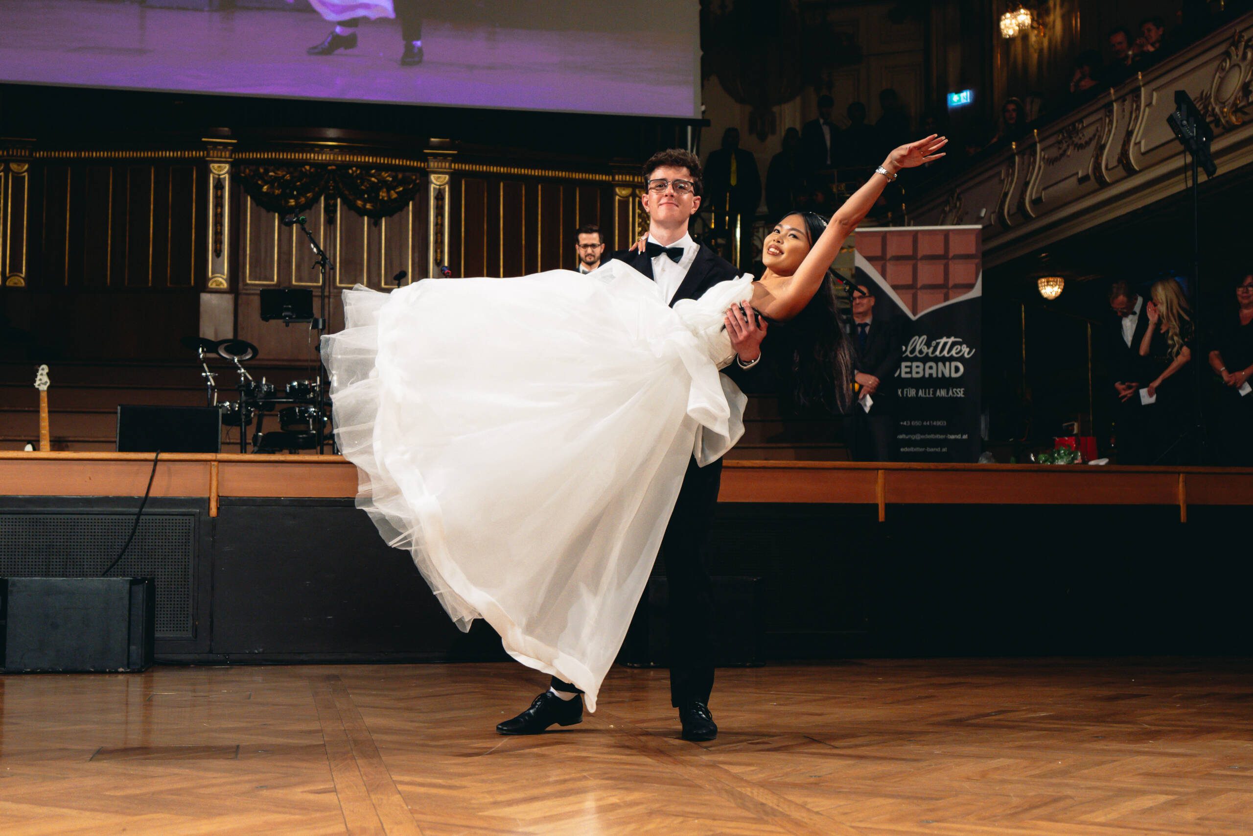 Ein Mann im Smoking hebt eine Frau in einem weißen Ballkleid hoch, während er in einem großen Ballsaal tanzt, während das Publikum von Balkonen und von der Seite zusieht.
