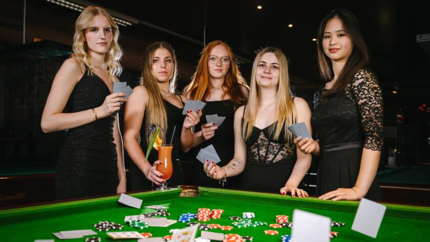 Fünf Frauen in eleganten schwarzen Kleidern stehen um einen grünen Pokertisch, auf dem Karten und Pokerchips verstreut sind, halten Spielkarten und Getränke in der Hand und schauen in einem schwach beleuchteten Raum in die Kamera.